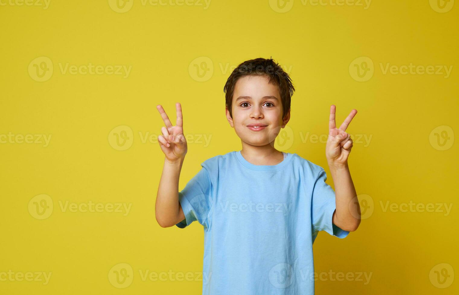 adorable garçon posant dans le caméra plus de Jaune Contexte avec copie espace. enfant faire des gestes la victoire avec les doigts photo