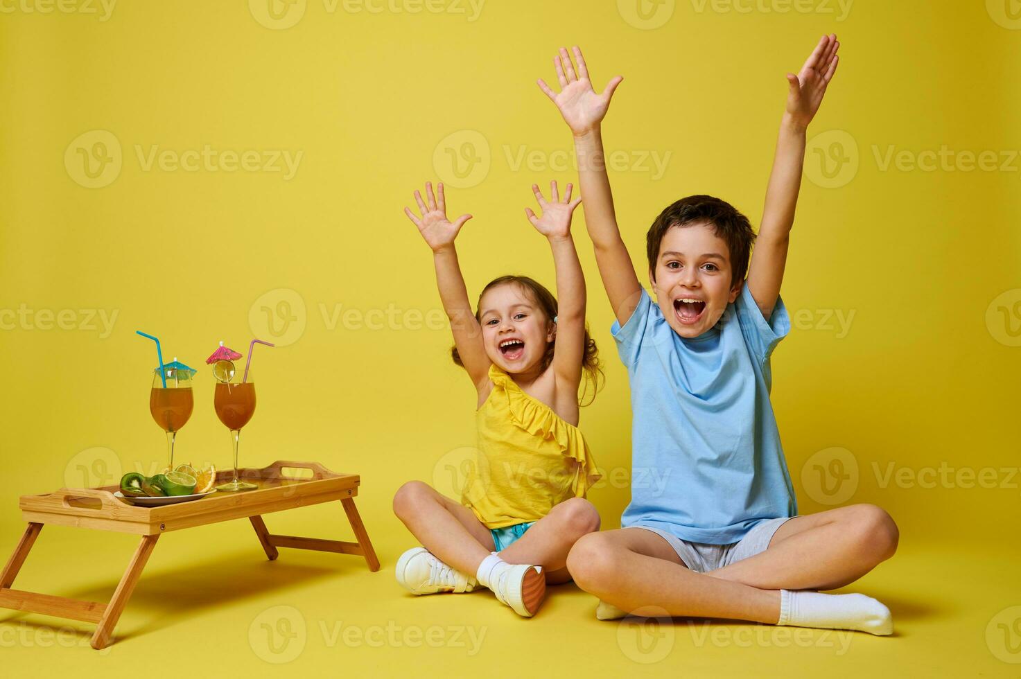 deux adorable les enfants séance dans lotus position près une en bois portion plateau avec des cocktails et assiette avec tranché des fruits et élevage bras en haut, en riant et exprimer bonheur photo