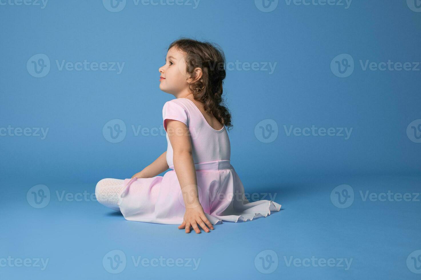 arrière vue de une repos préscolaire fille ballerine habillé dans rose uniforme après dansant, séance sur bleu Contexte photo