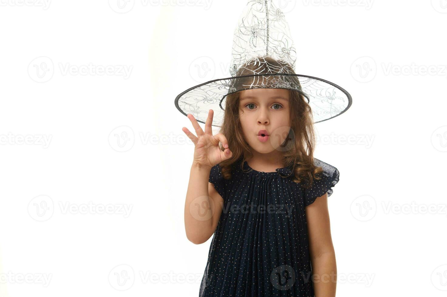 isolé taille longueur portrait de jolie peu sorcière fille portant une sorcier chapeau et habillé dans élégant carnaval robe, faire des gestes, montrant D'accord signe avec des doigts. Halloween concept sur blanc Contexte photo