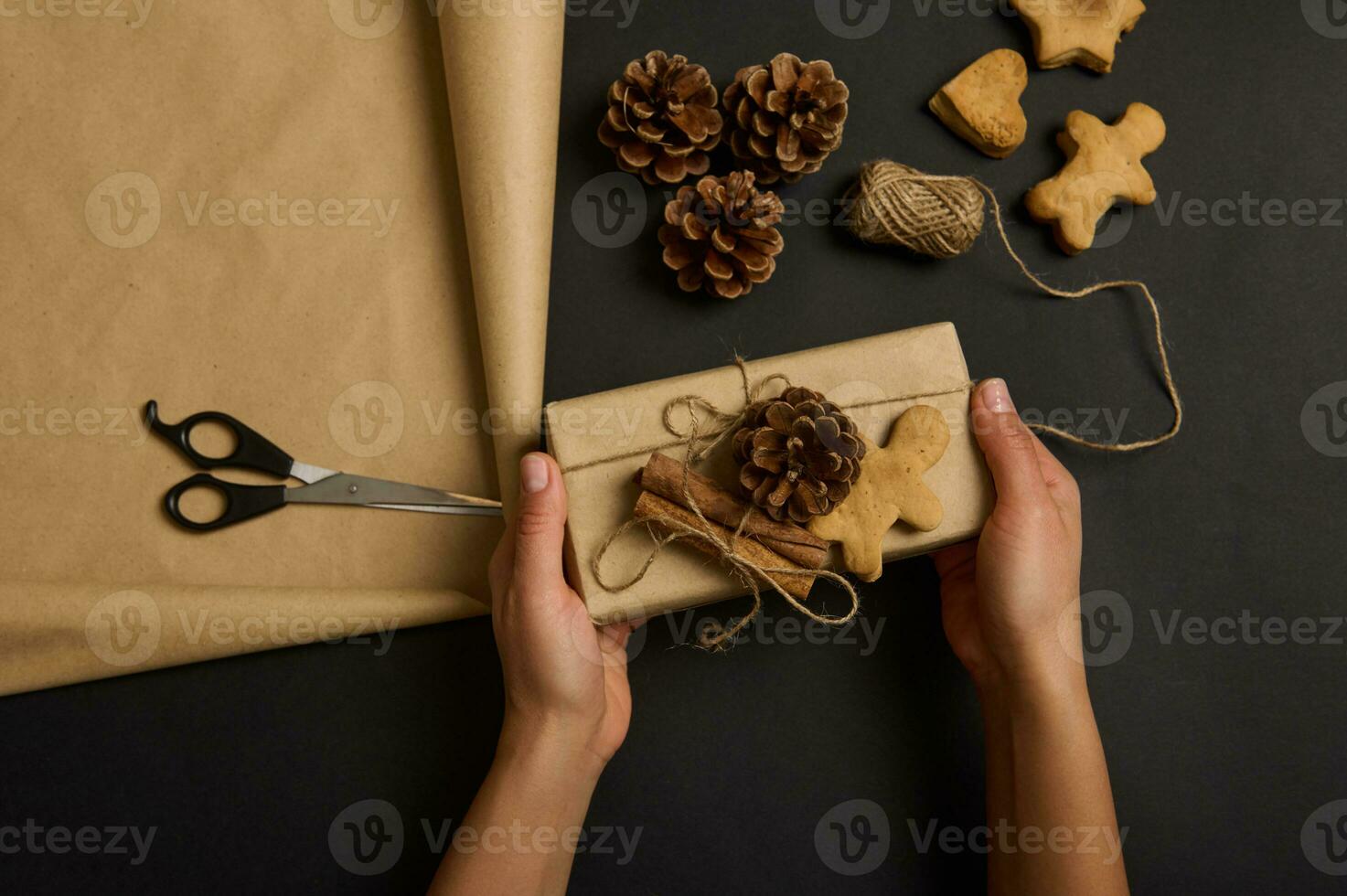 mains en portant Fait main Noël cadeau boîte enveloppé dans marron artisanat papier décoré avec pin cônes et cannelle mensonge sur une noir surface avec une pain d'épice homme et des biscuits, corde et ciseaux. plat allonger photo