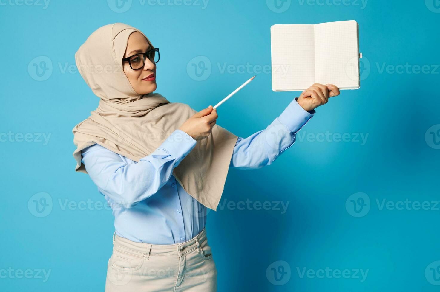 musulman femme dans hijab détient Vide livre et points à il avec stylo isolé sur bleu Contexte photo