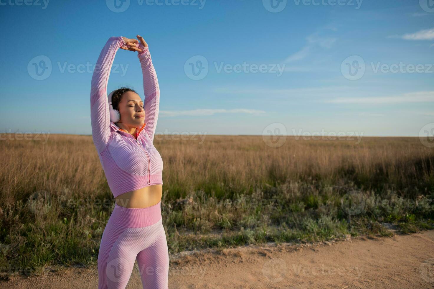 attrayant femelle coureur dans rose tenue de sport s'étire sa bras en haut tandis que permanent sur le steppe route avec fermé yeux profiter le magnifique le coucher du soleil tandis que le jogging photo