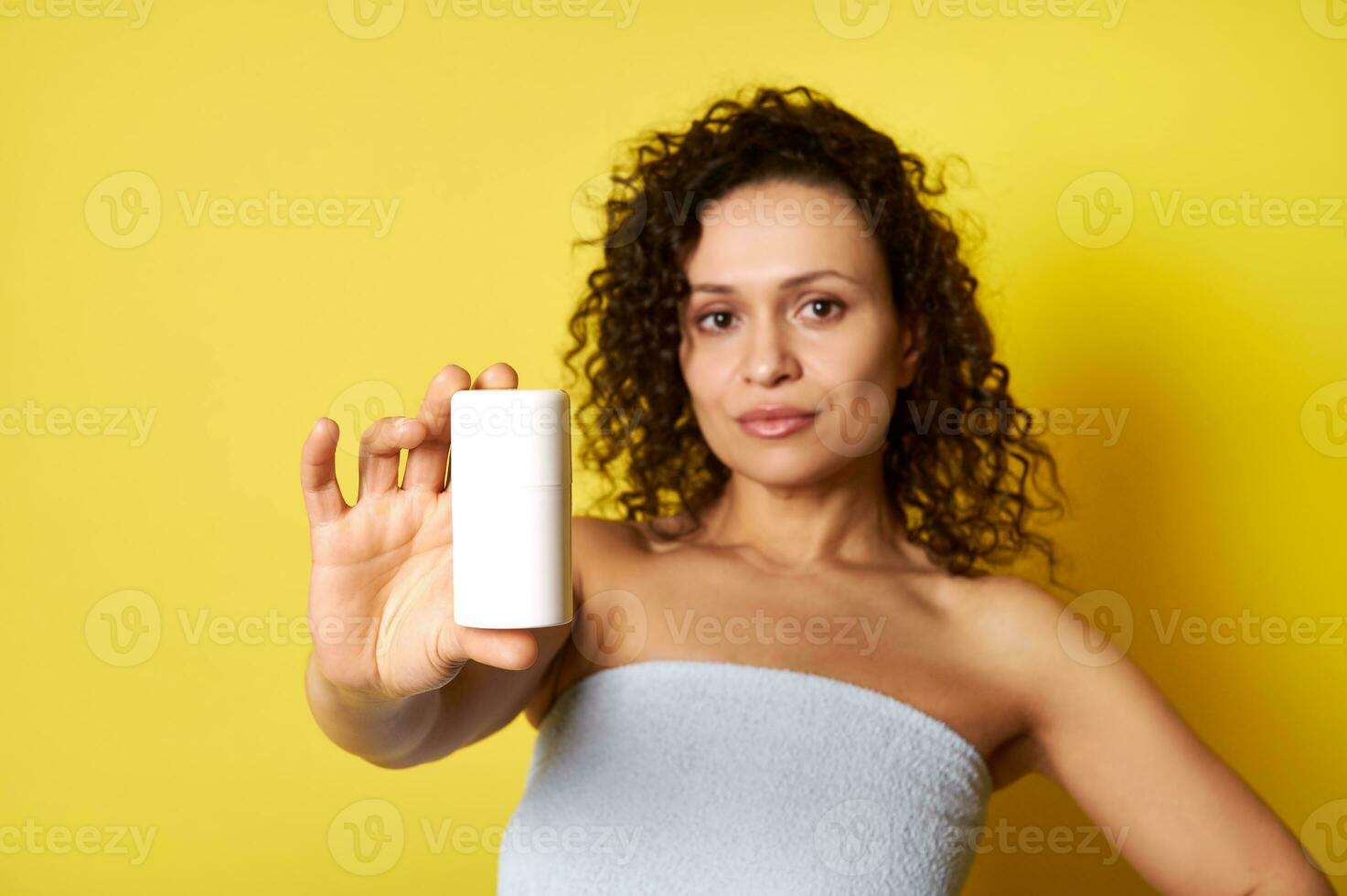 doux concentrer sur blanc récipient avec cosmétique produit dans main de magnifique moitié nu frisé femme enveloppé dans une baignoire serviette photo