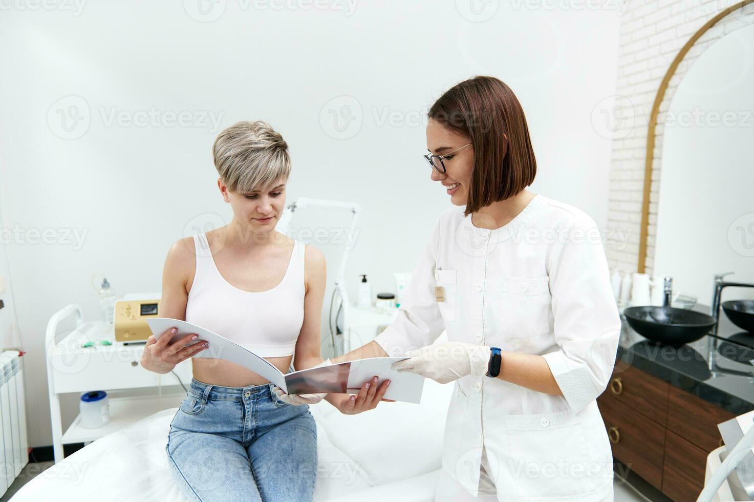 amical cosmétologue montrant une catalogue de des prix pour cosmétique procédures à une blond client séance sur un fauteuil dans une beauté parloir pendant une consultation à une beauté spa clinique photo
