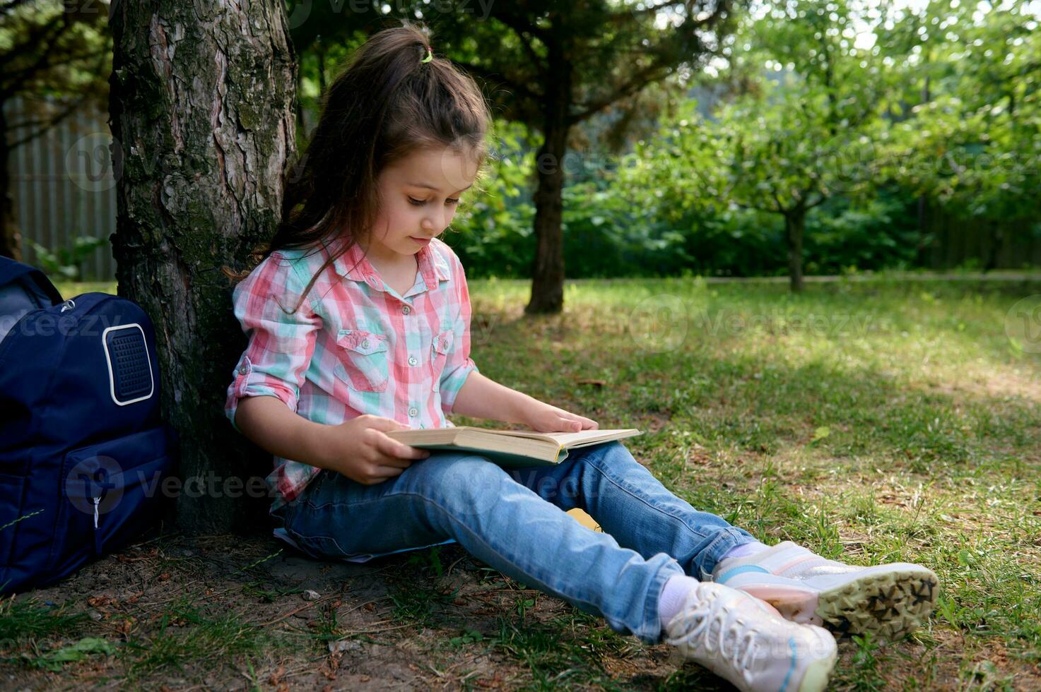 peu école fille lit une livre dans le parc, séance en dessous de arbre avec sa sac à dos. enfants. érudition. éducation. mode de vie photo