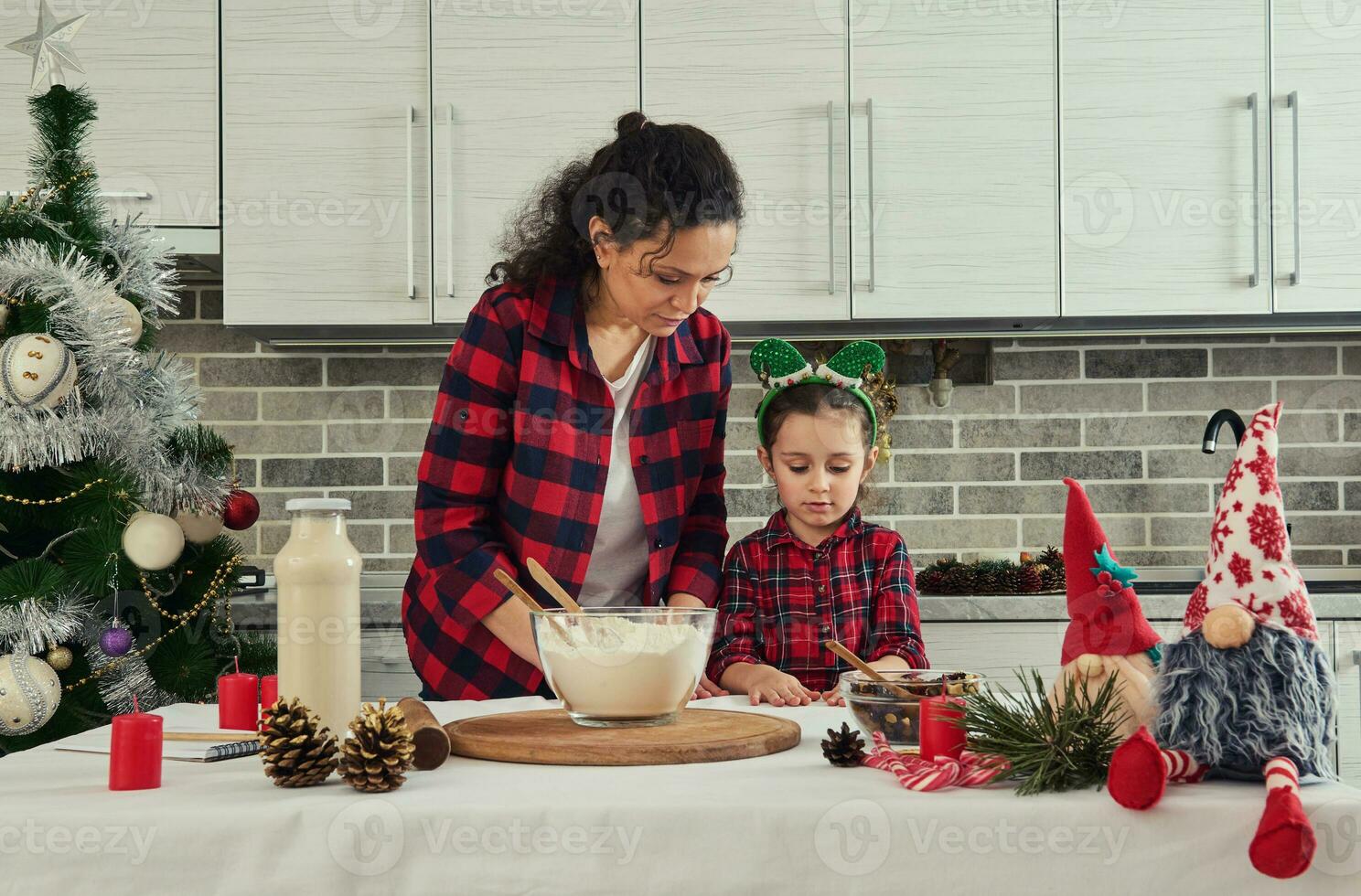 content maman avec sa adorable bébé fille fille cuisine ensemble à Accueil cuisine. Ingrédients pour Noël gâteau sur le table suivant à Fait main scandinave gnomes, rouge bougies, pin cônes, bonbons cannes photo