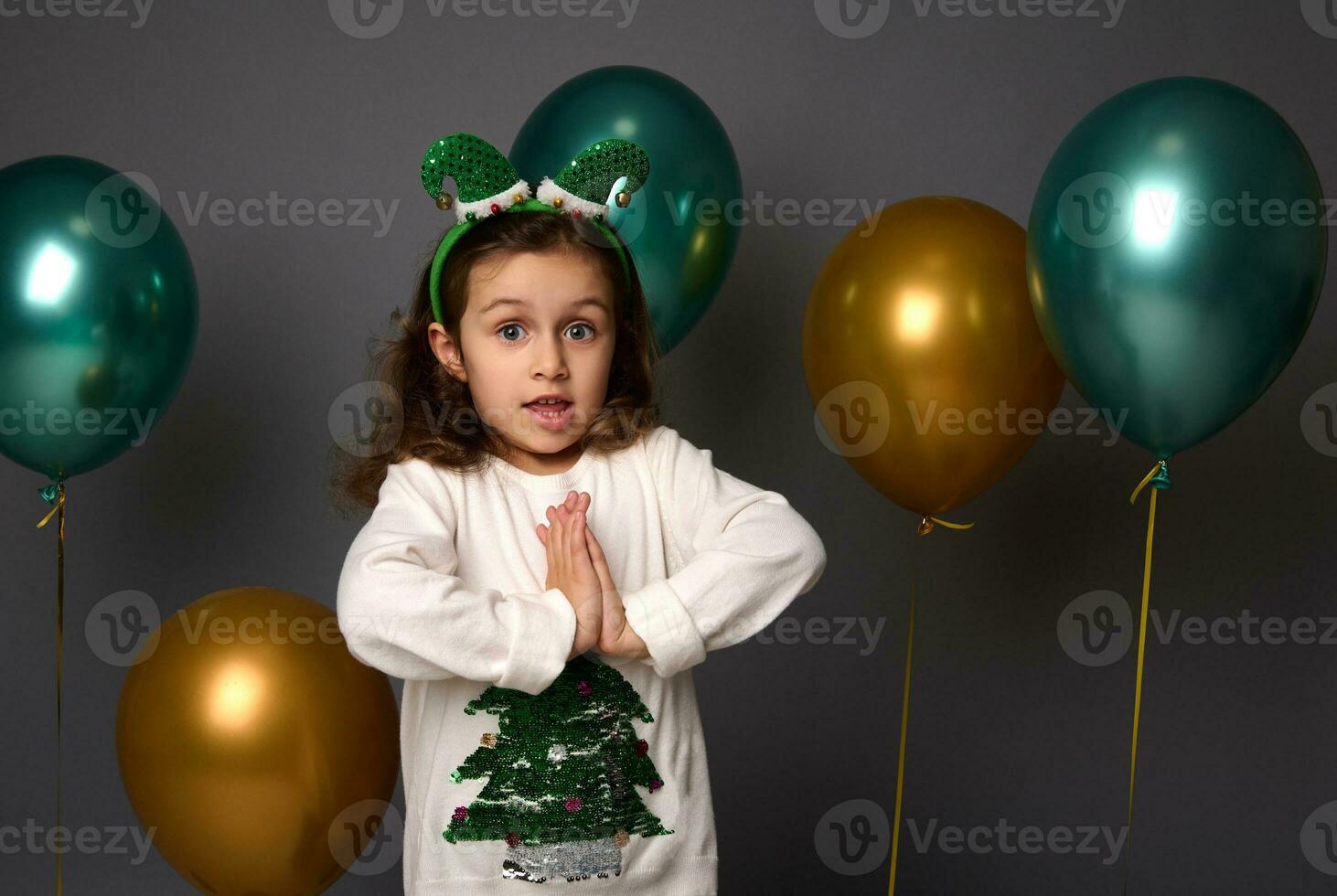 magnifique peu fille dans Noël chandail et elfe cerceau pose avec mains paume à palmier, regards à caméra, isolé plus de gris Contexte avec d'or métallique vert air des ballons avec copie espace pour un d photo