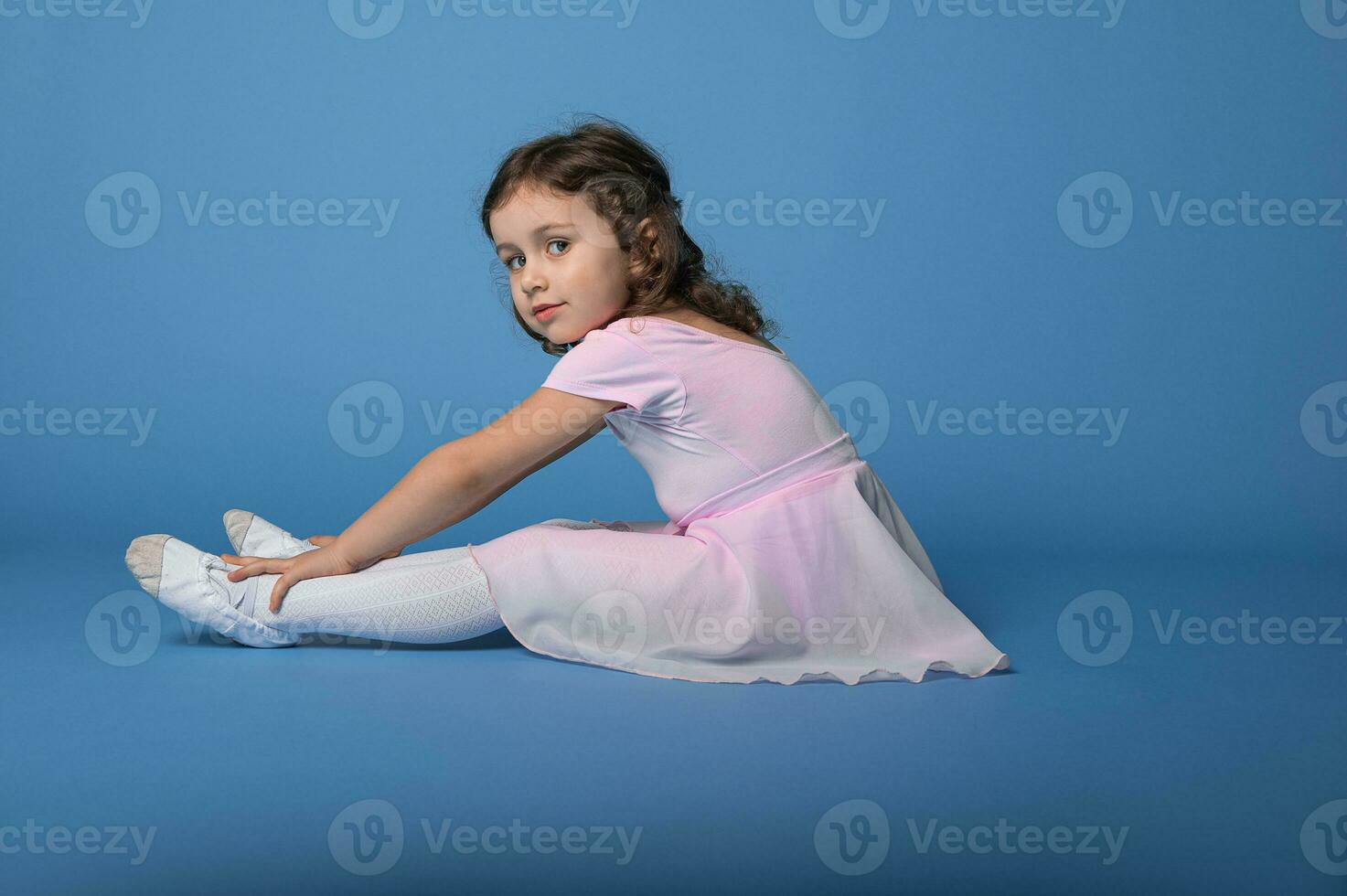 côté portrait de une peu ballerine dans rose robe performant élongation jambes, séance plus de bleu Contexte photo