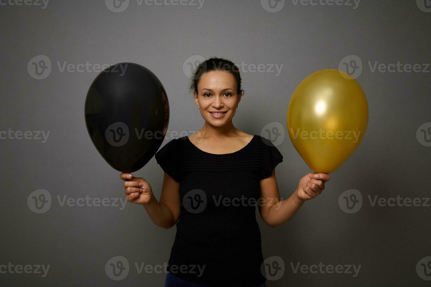 de bonne humeur africain américain femme habillé dans noir sourit à la recherche à caméra et pose contre gris Contexte avec une or et noir air des ballons dans main. noir Vendredi concept avec copie un d espace photo