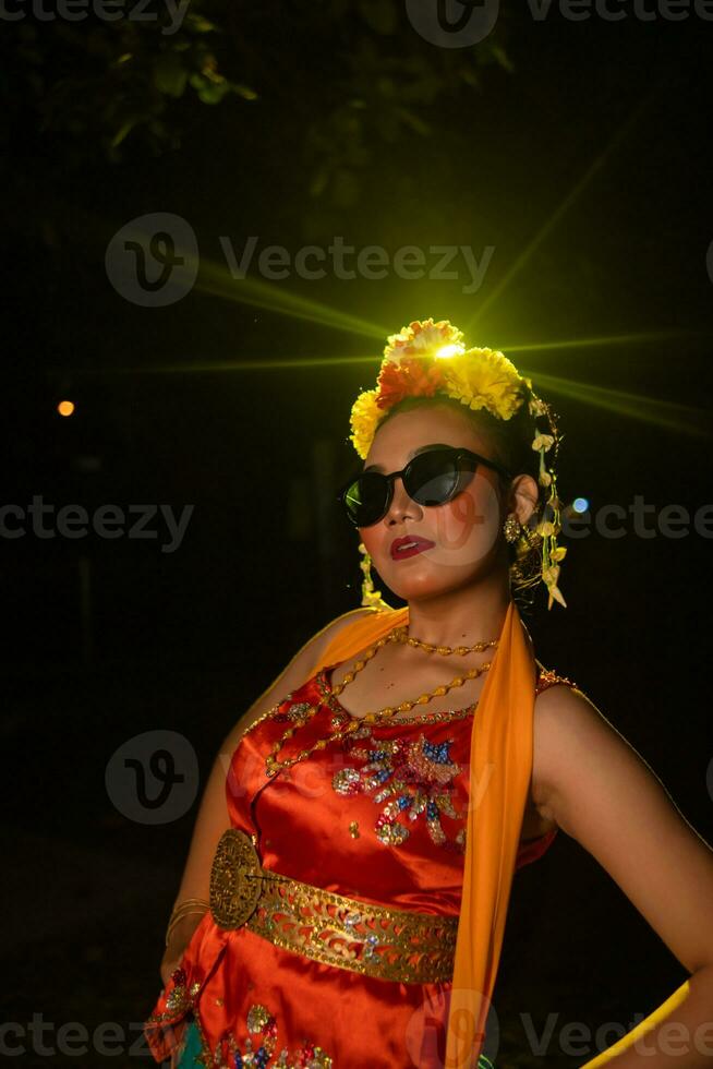 une sundanais Danseur pose dans de face de une brillant lumière tandis que portant un Orange costume avec des lunettes de soleil et fleurs sur sa tête photo