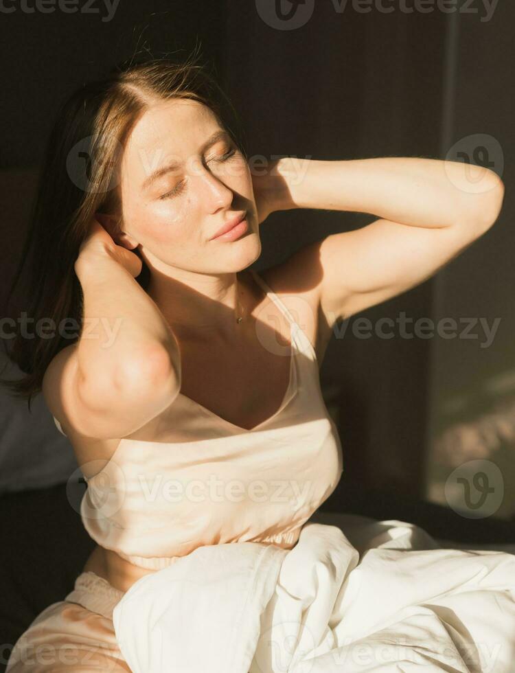 portrait femme veille en haut dans sa lit, elle est souriant et élongation. content Jeune femme salue Nouveau ensoleillé journée photo