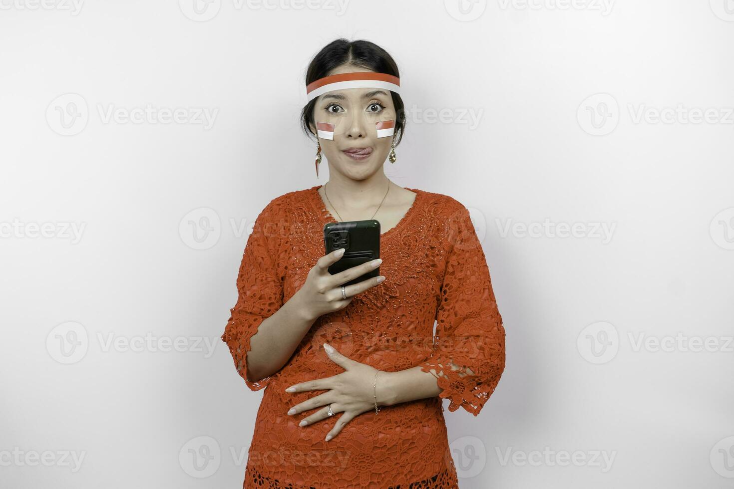 une réfléchi asiatique femme portant rouge kebaya et Bandeau, en portant sa téléphone et estomac, en pensant à propos quoi à manger ou commande, isolé par blanc Contexte. l'indonésie indépendance journée photo