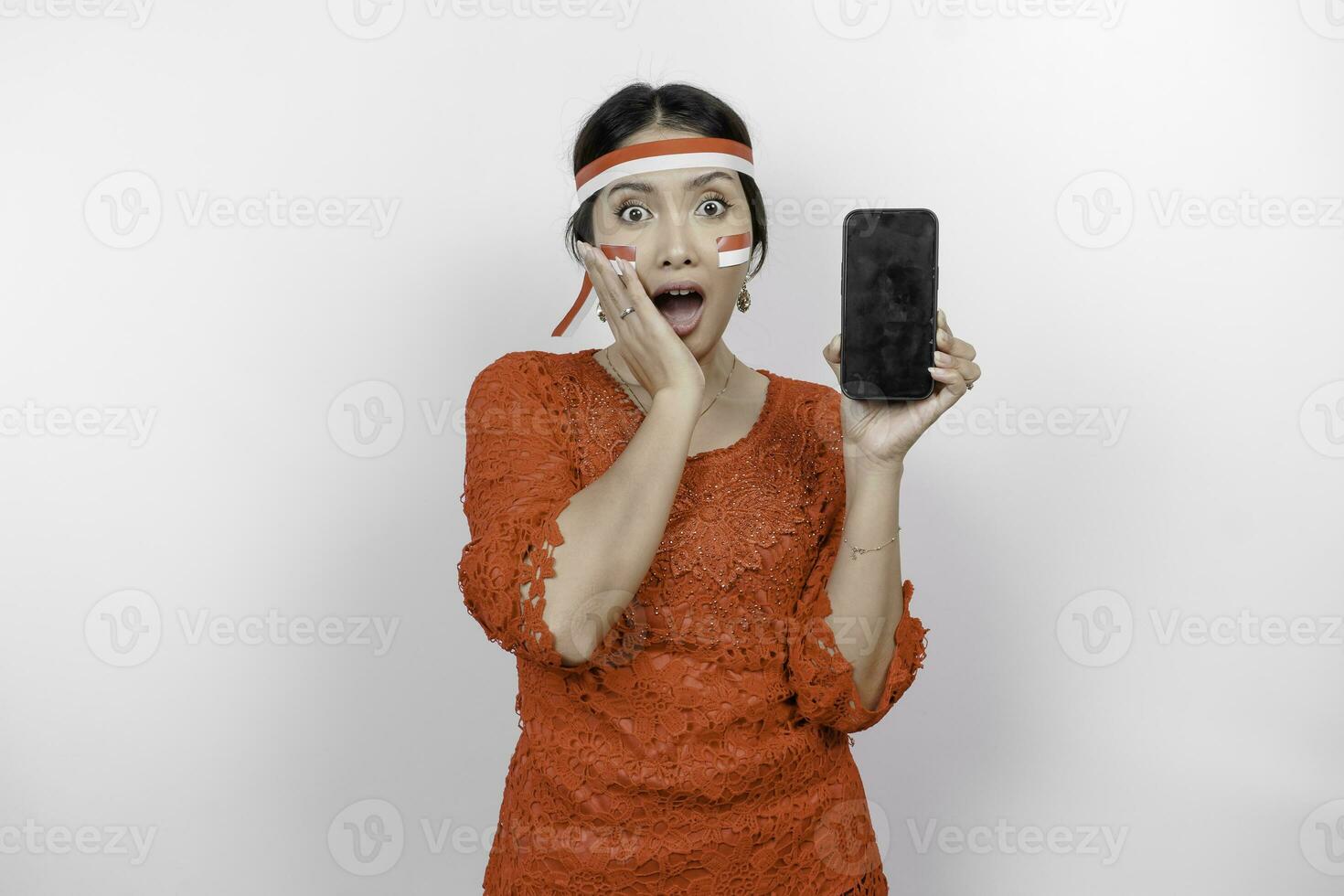 une portrait de une sous le choc asiatique femme portant rouge kebaya et Bandeau, montrant sa téléphone tandis que sa bouche large ouvrir, isolé par blanc Contexte. l'indonésie indépendance journée concept photo