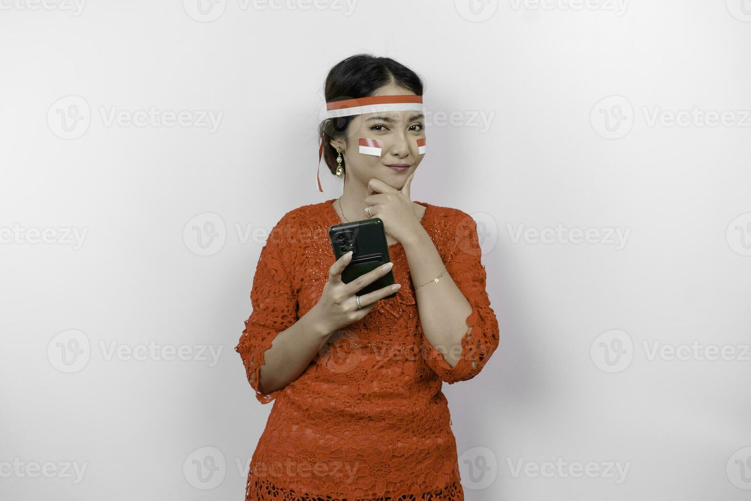 une intelligent Jeune femme portant Bandeau et rouge kebaya est à la recherche de côté à un idée sur copie espace tandis que en portant sa téléphone, isolé par blanc Contexte. l'indonésie indépendance journée. photo