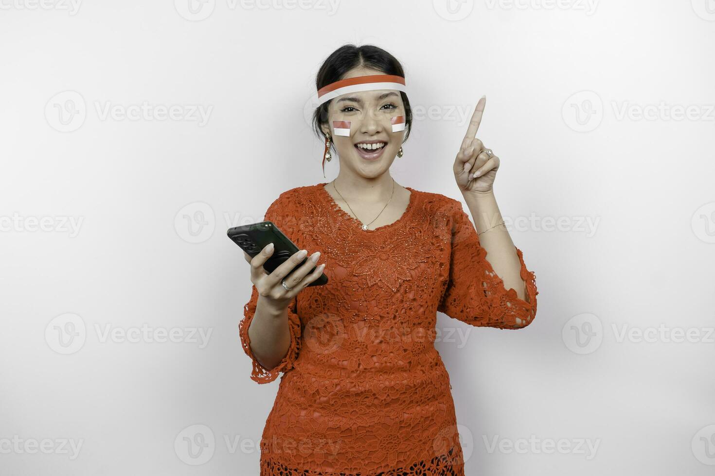 une content asiatique femme portant rouge kebaya et Bandeau, en portant sa téléphone, et montrer du doigt copie espace sur Haut de son, isolé par blanc Contexte. l'indonésie indépendance journée photo