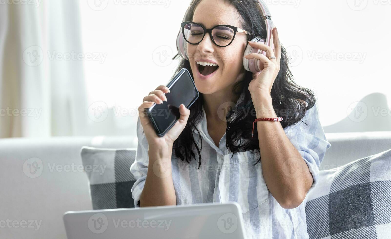 excité femme en utilisant téléphone portable comme microphone, séance sur  canapé et écoute à la musique par sans fil écouteurs de portable. 27486057  Photo de stock chez Vecteezy