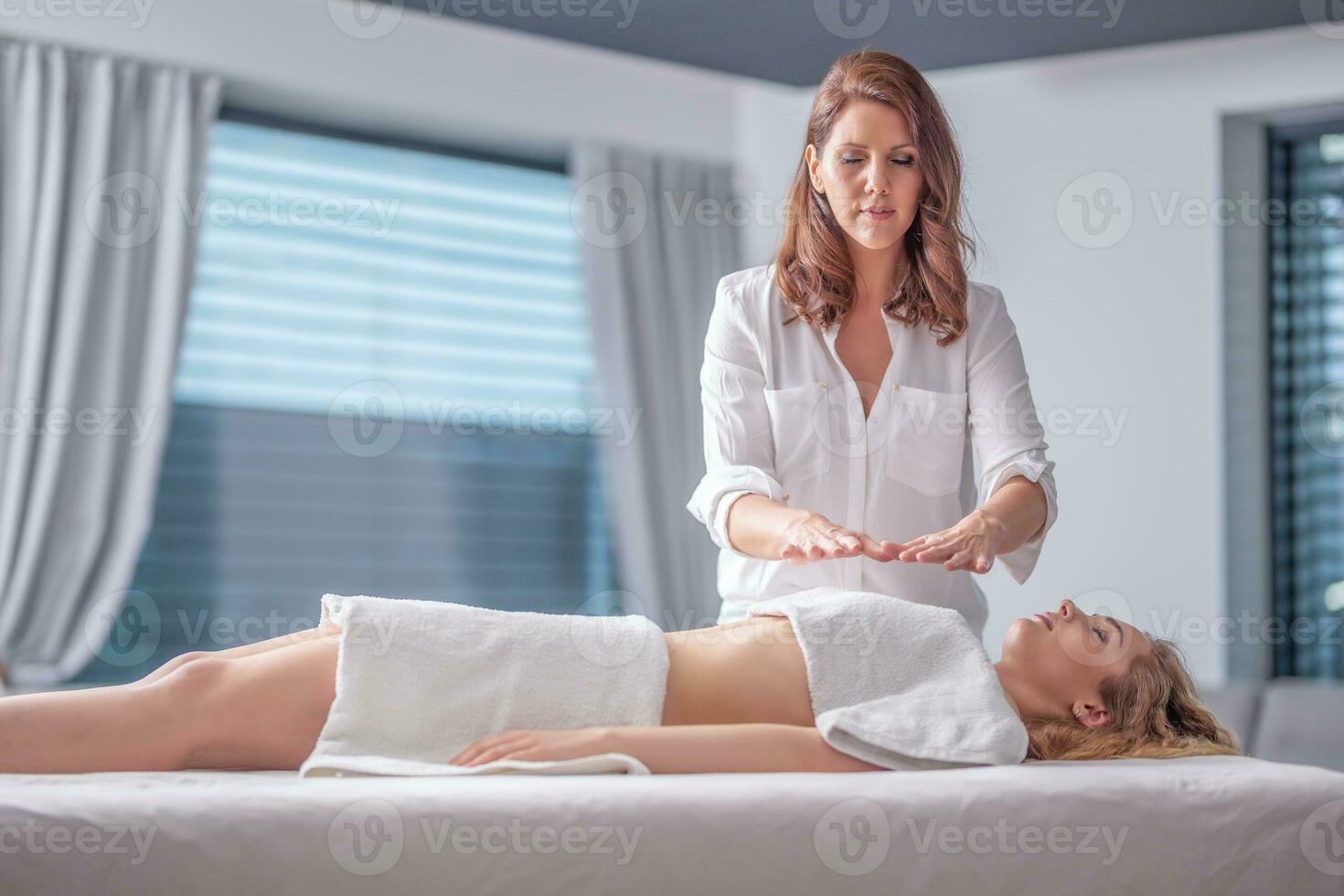 détendu Jeune femme couvert par les serviettes ayant reiki guérison avec une se concentrer thérapeute avec fermé yeux photo