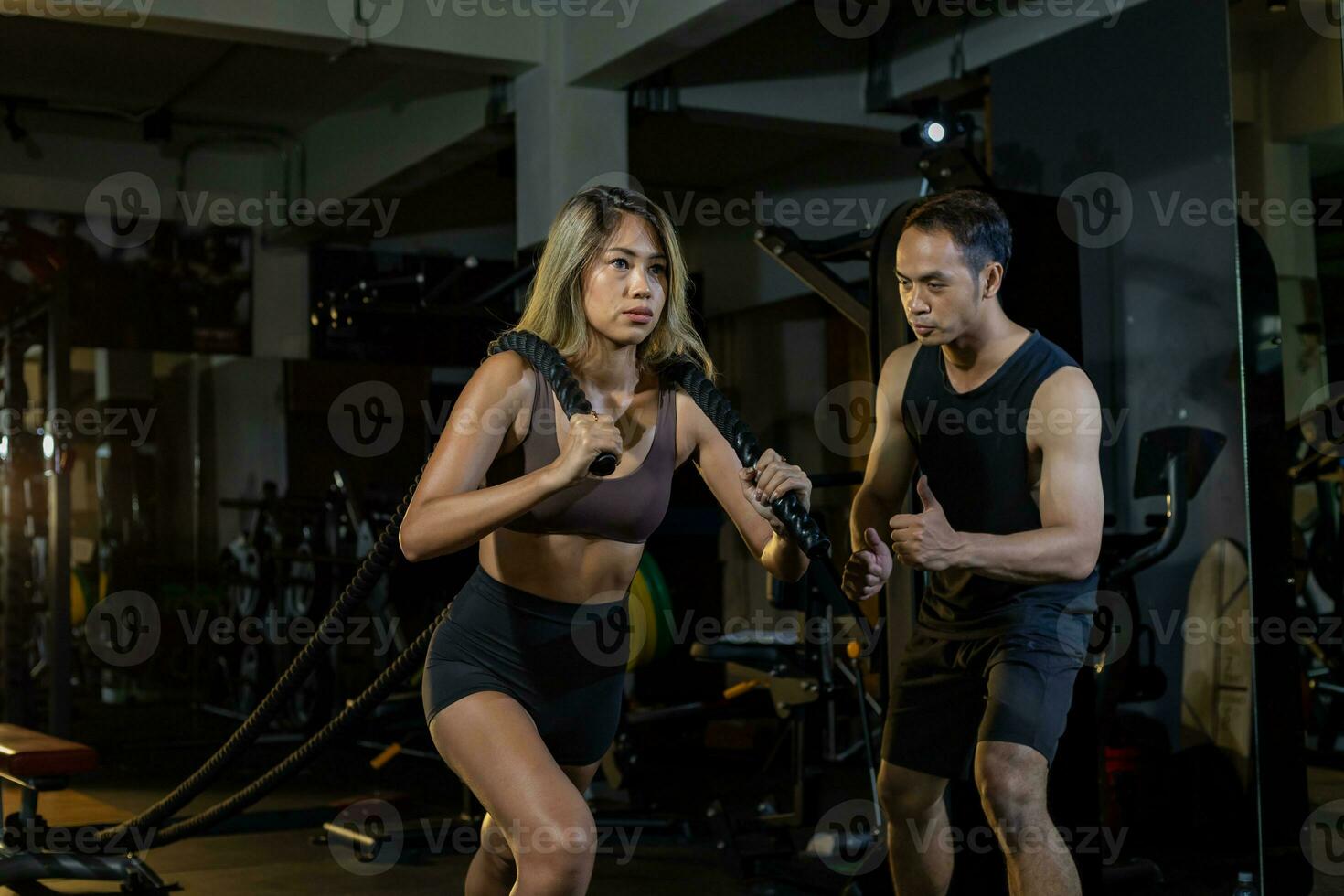 asiatique femme avec fort et musclé corps formation sur coeur poids formation par corde tirant avec sa justificatif entraîneur encourageant à l'intérieur Gym pour avance exercice et faire des exercices concept photo