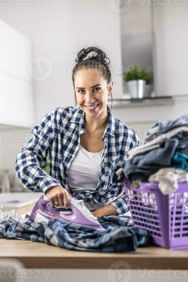 femme dans bien ambiance sourit et fers vêtements avec une violet le fer et vêtements dans une violet Plastique panier photo