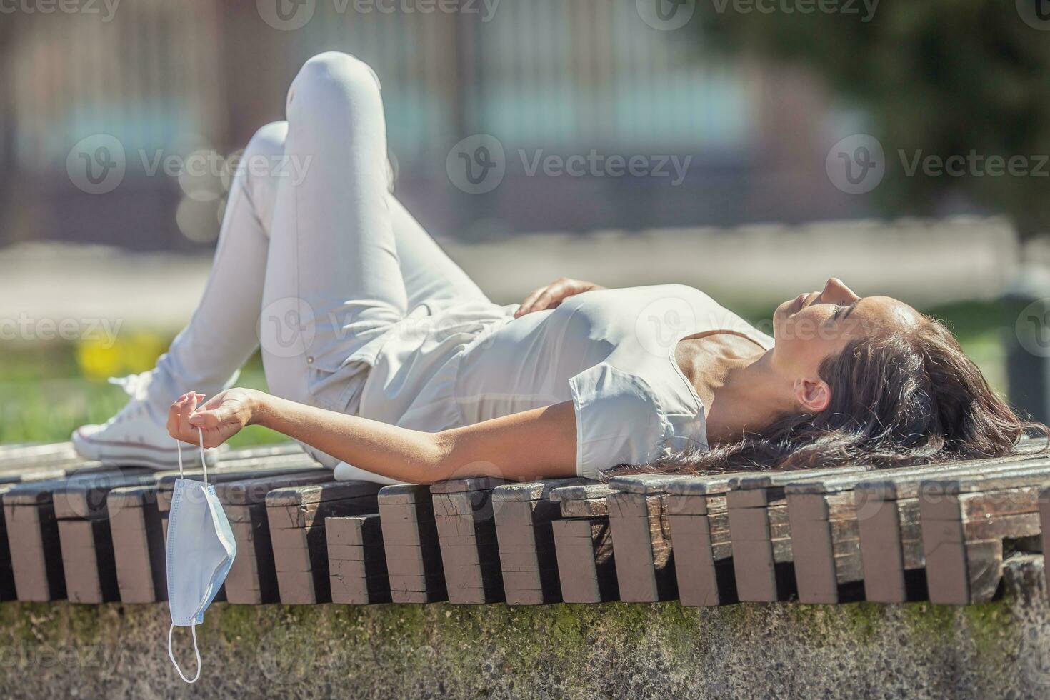 Jeune femme mensonges vers le bas sur une banc dans le ville, bain de soleil, en portant visage masque par une sangle dans sa main photo