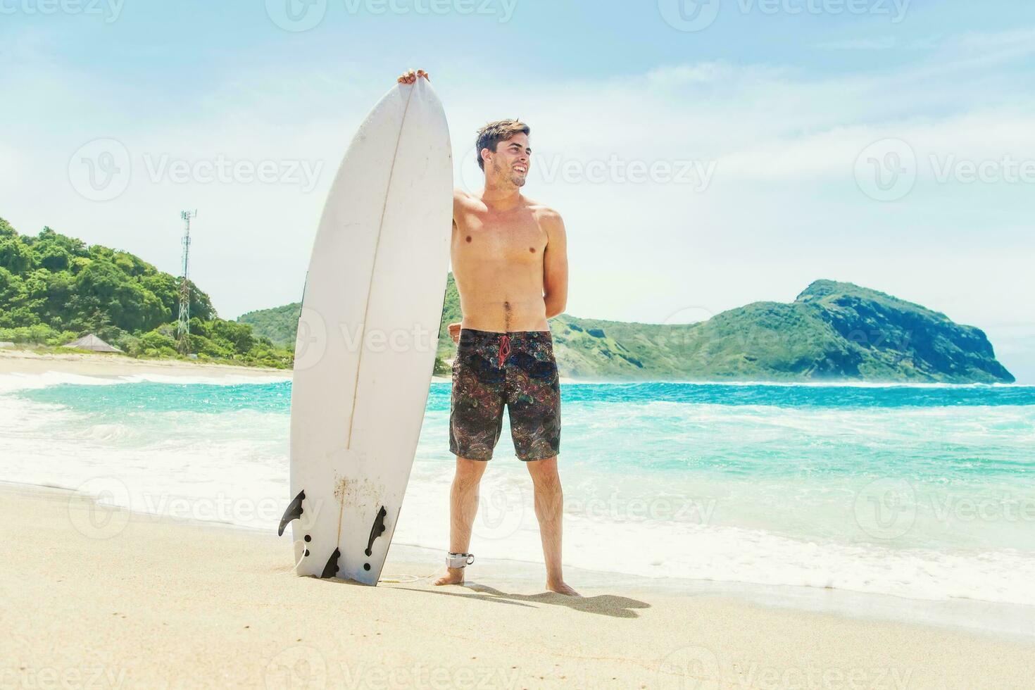 une homme permanent sur le plage en portant une planche de surf photo