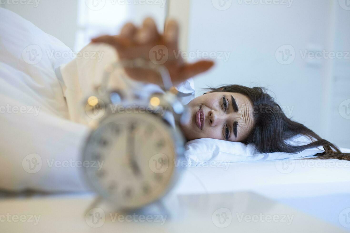 femme mensonge dans lit tournant de un alarme l'horloge dans le Matin à 5h du matin. main se tourne de le alarme l'horloge veille en haut à matin, fille se tourne de le alarme l'horloge veille en haut dans le Matin de une appel. photo