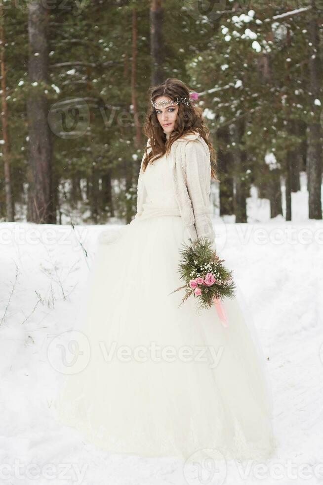 magnifique la mariée dans une blanc robe avec une bouquet dans une couvert de neige hiver forêt. portrait de le la mariée dans la nature photo