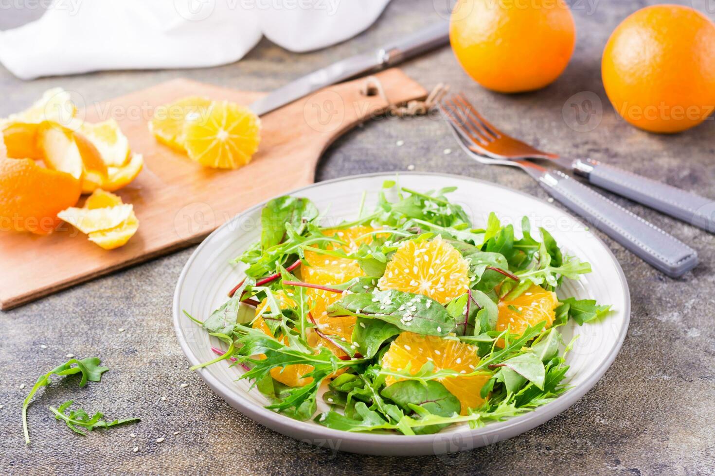 régime végétarien vitamine salade de Orange tranches et une mélanger de roquette, bettes et mizun feuilles sur une assiette et une Coupe planche avec pelé Orange sur le table photo