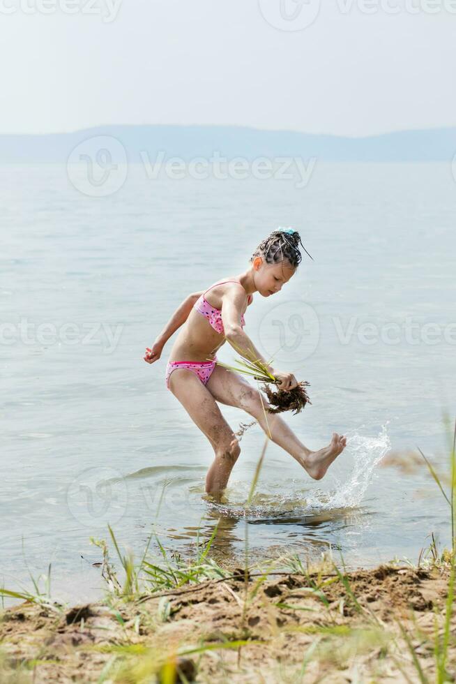 une adolescent fille dans une baignade costume fanns sa jambe avec une balai de herbe imitant une baignoire procédures sur le ensoleillé banque de le rivière dans été. vacances Activités pour les enfants photo