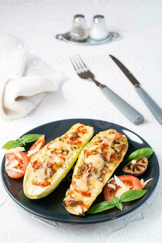 prêt à manger cuit Zucchini moitiés rempli avec fromage et tomate et basilic feuilles et coutellerie sur une noir assiette sur une blanc tableau. légume menu, en bonne santé aliments. verticale vue photo