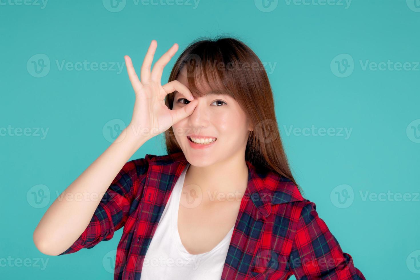 belle jeune femme asiatique heureuse porte un geste d'été de voyage signe ok avec la main sur l'œil ayant une vision isolée sur fond bleu, expression d'une adolescente asiatique avec une expression joyeuse et amusante pendant le voyage. photo