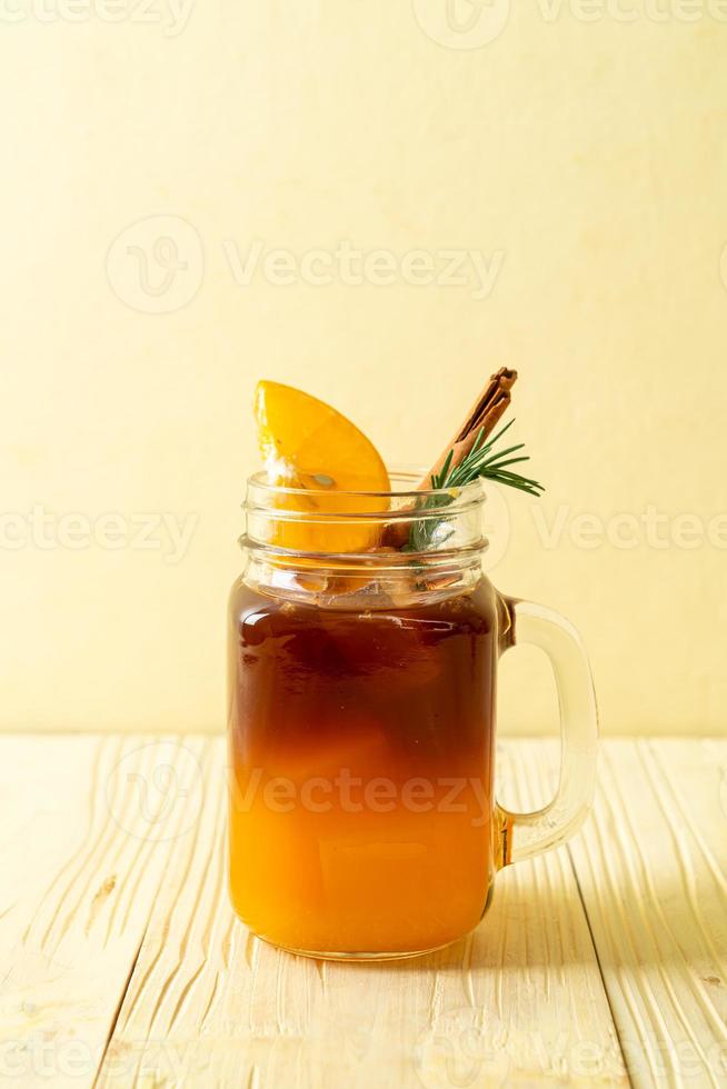 un verre de café noir americano glacé et une couche de jus d'orange et de citron décoré de romarin et de cannelle sur fond de bois photo