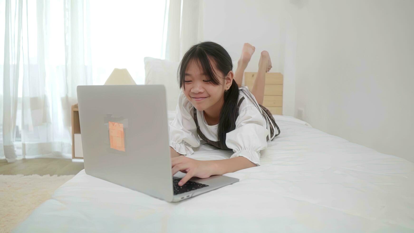 content peu fille enfant. mignonne peu fille mensonge sur le sien estomac une lit et en train de regarder quelque chose intéressant sur une portable. photo