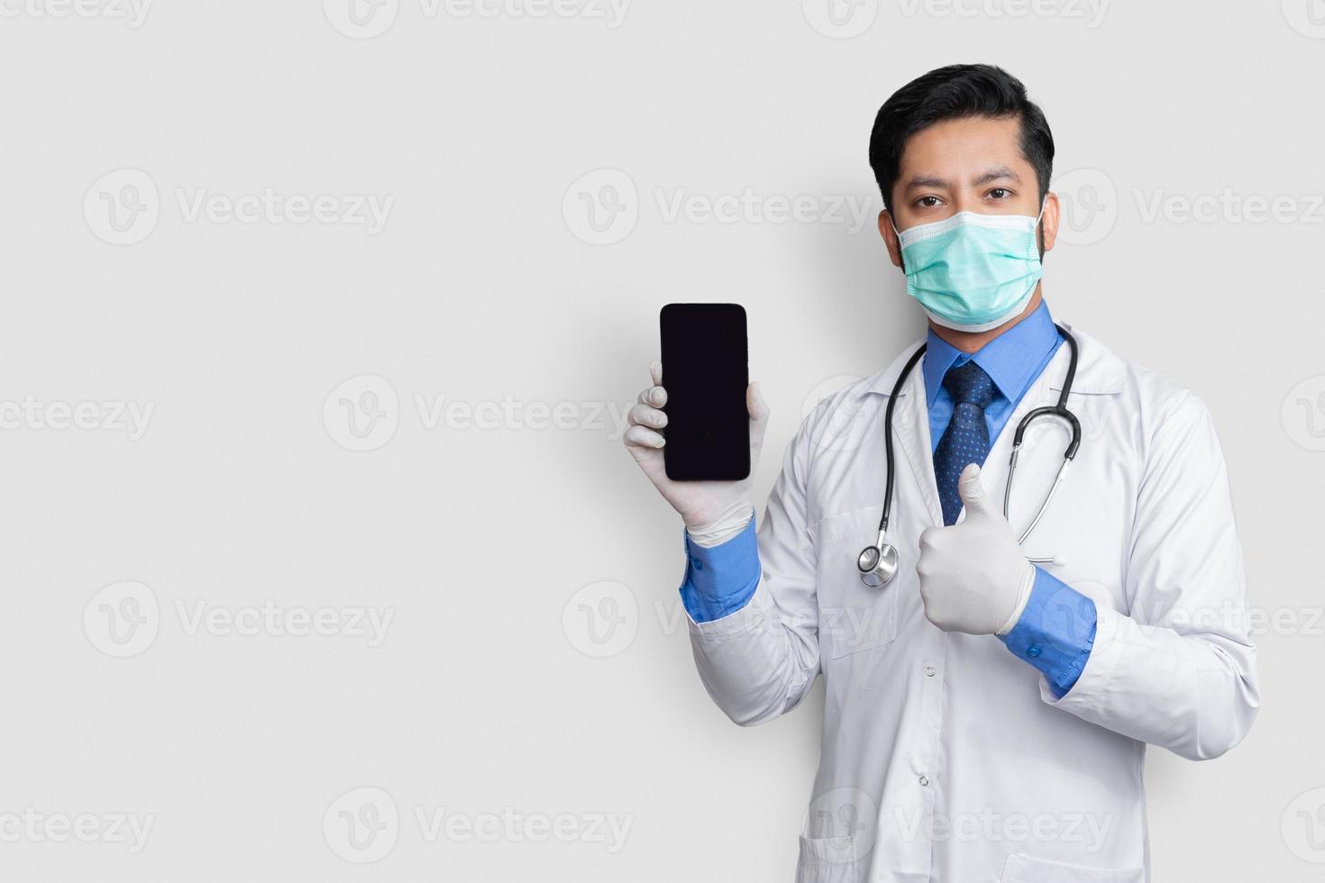 docteur tenant le téléphone montrant le pouce vers le haut isolé. pakistanais homme médecin technologie médecine à la maison. écran clair du téléphone. photo