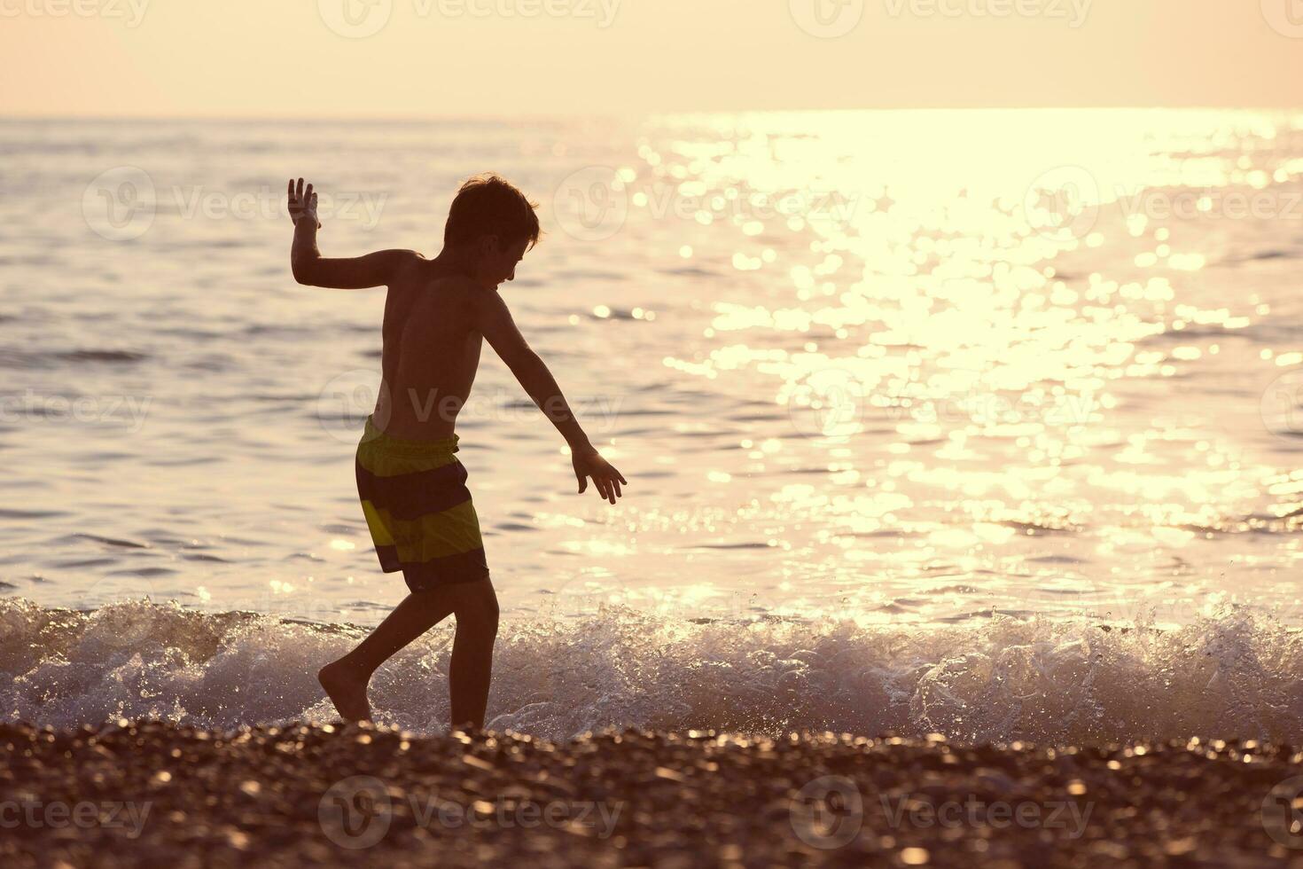 silhouette de une garçon par le mer à le coucher du soleil. il court le long de le rive, pièces avec le vague. photo