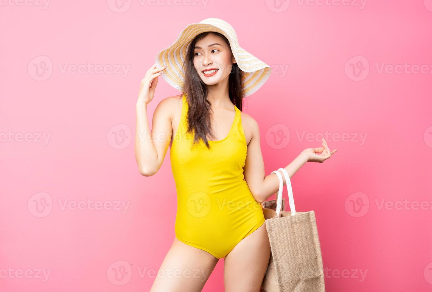 belle jeune femme asiatique en maillot de bain jaune, sac et chapeau à larges bords posant sur fond rose photo