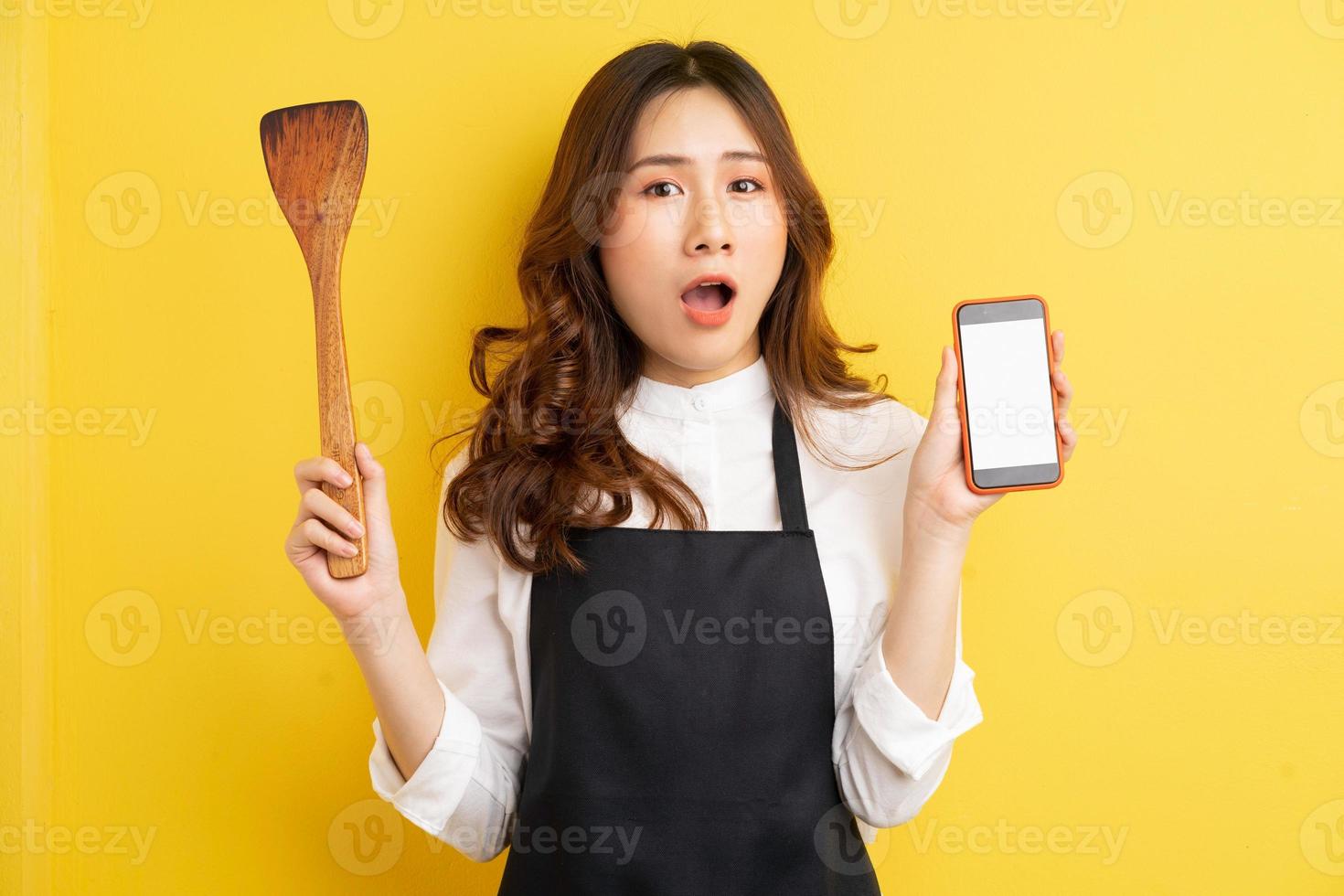 belle femme au foyer tenant une cuillère à riz et un téléphone avec une heureuse surprise isolée sur fond jaune photo