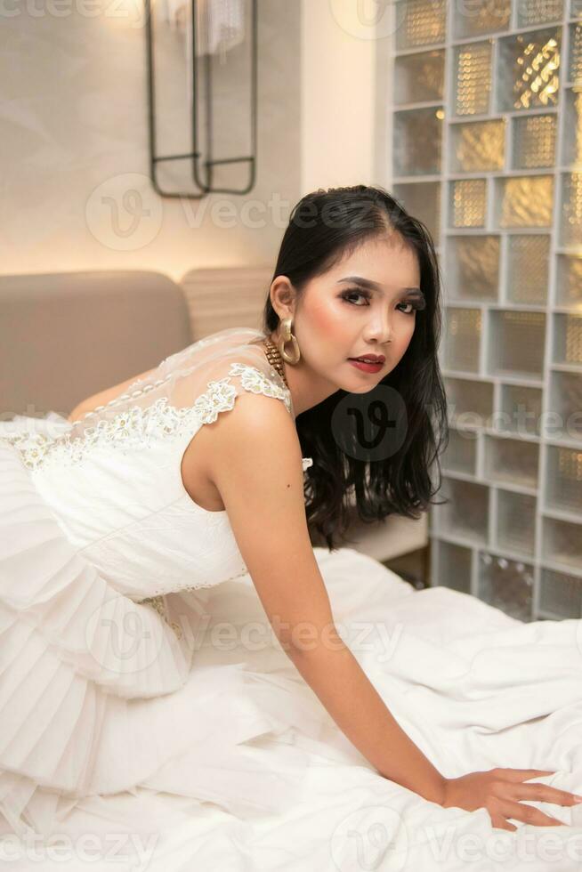 un asiatique femme dans une blanc robe est posant sexy avec une visage cette regards corné sur une blanc lit photo