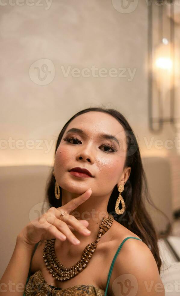 un asiatique femme avec une or Collier et des boucles d'oreilles est posant magnifiquement dans une Hôtel pièce avant dîner photo