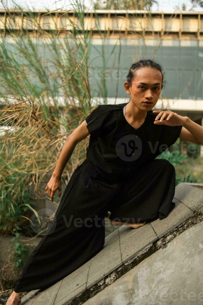 un asiatique homme pose sur une en pente Roche tandis que portant noir autour un abandonné bâtiment photo