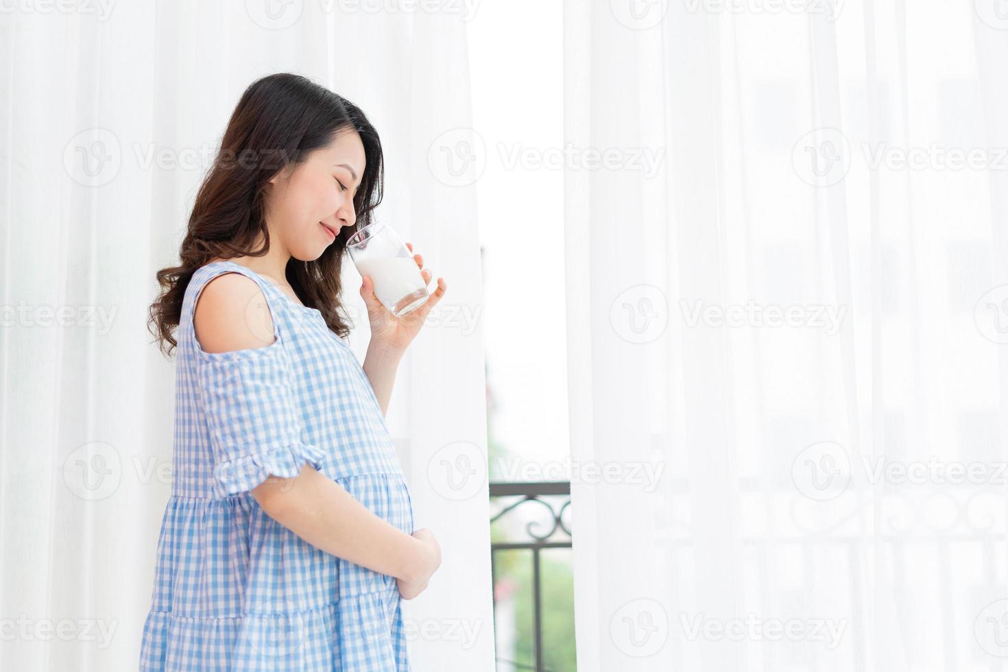 belle femme tenant son ventre et tenant une tasse de lait se tient près de la fenêtre photo