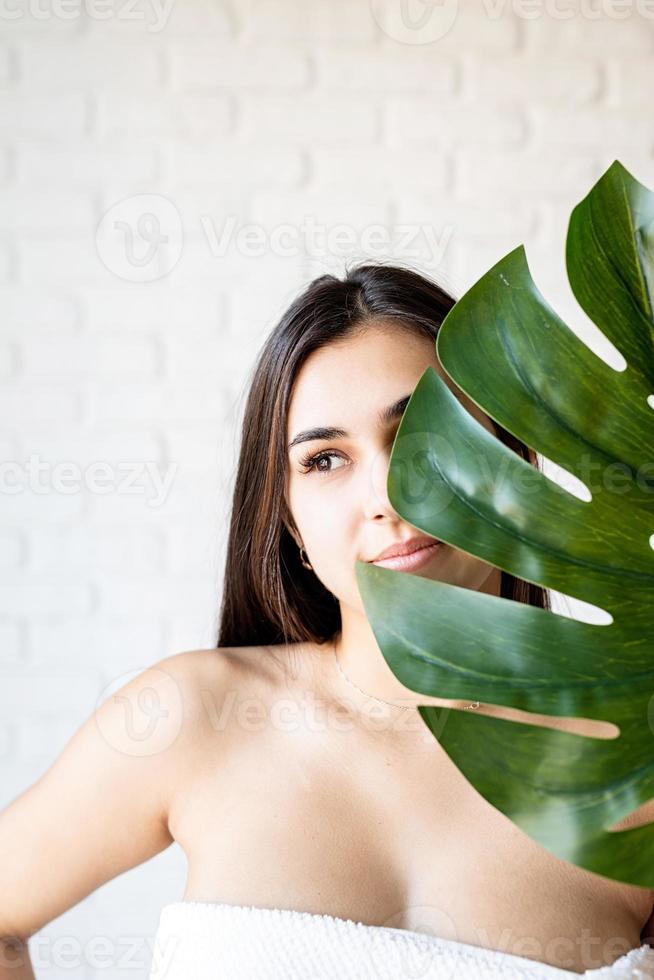 heureuse belle femme portant des serviettes de bain tenant une feuille de monstera verte devant son visage photo