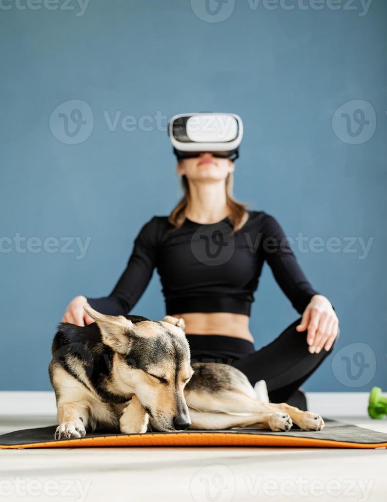 Jeune femme en vêtements de sport portant des lunettes de réalité virtuelle assis sur un tapis de fitness avec chien photo