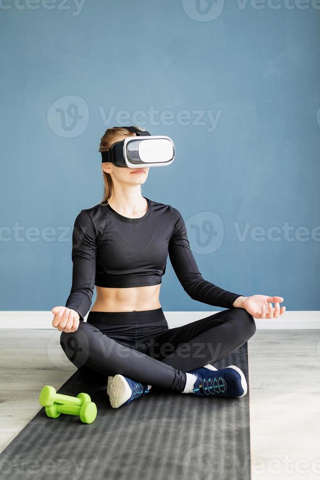 Jeune femme blonde en vêtements de sport portant des lunettes de réalité virtuelle méditant sur un tapis de fitness photo