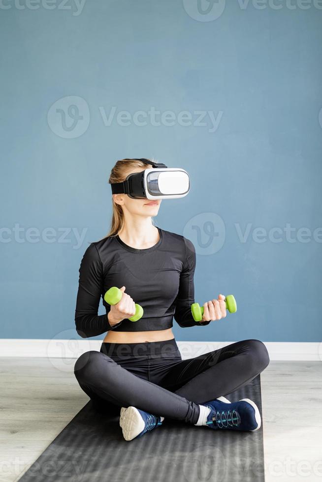Jeune femme blonde en vêtements de sport portant des lunettes de réalité virtuelle assis sur un tapis de fitness photo