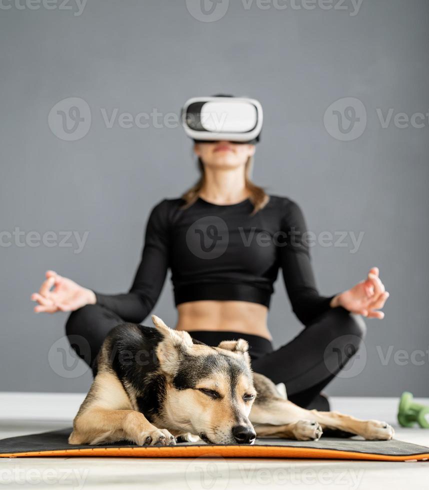 Jeune femme en vêtements de sport portant des lunettes de réalité virtuelle assis sur un tapis de fitness avec chien photo