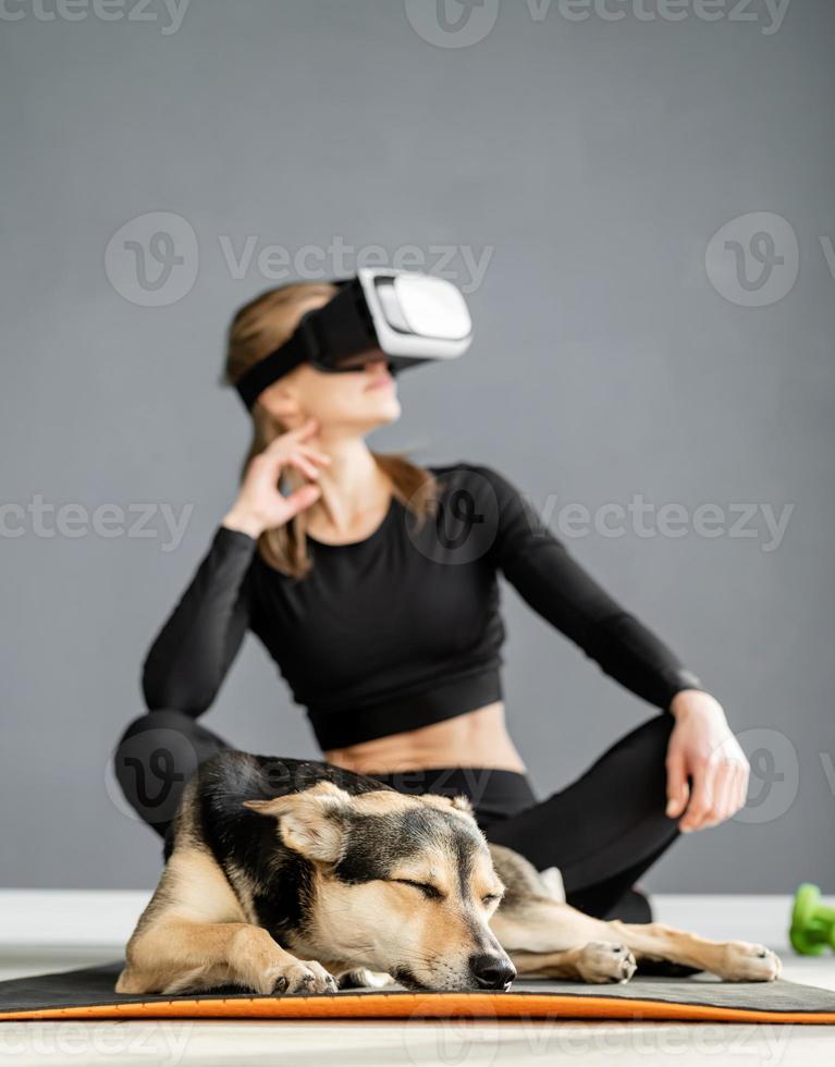 Jeune femme en vêtements de sport portant des lunettes de réalité virtuelle assis sur un tapis de fitness avec chien photo