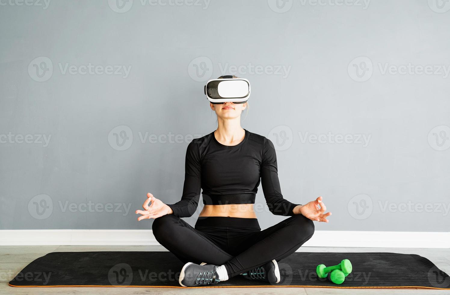 Jeune femme blonde en vêtements de sport portant des lunettes de réalité virtuelle méditant sur un tapis de fitness photo