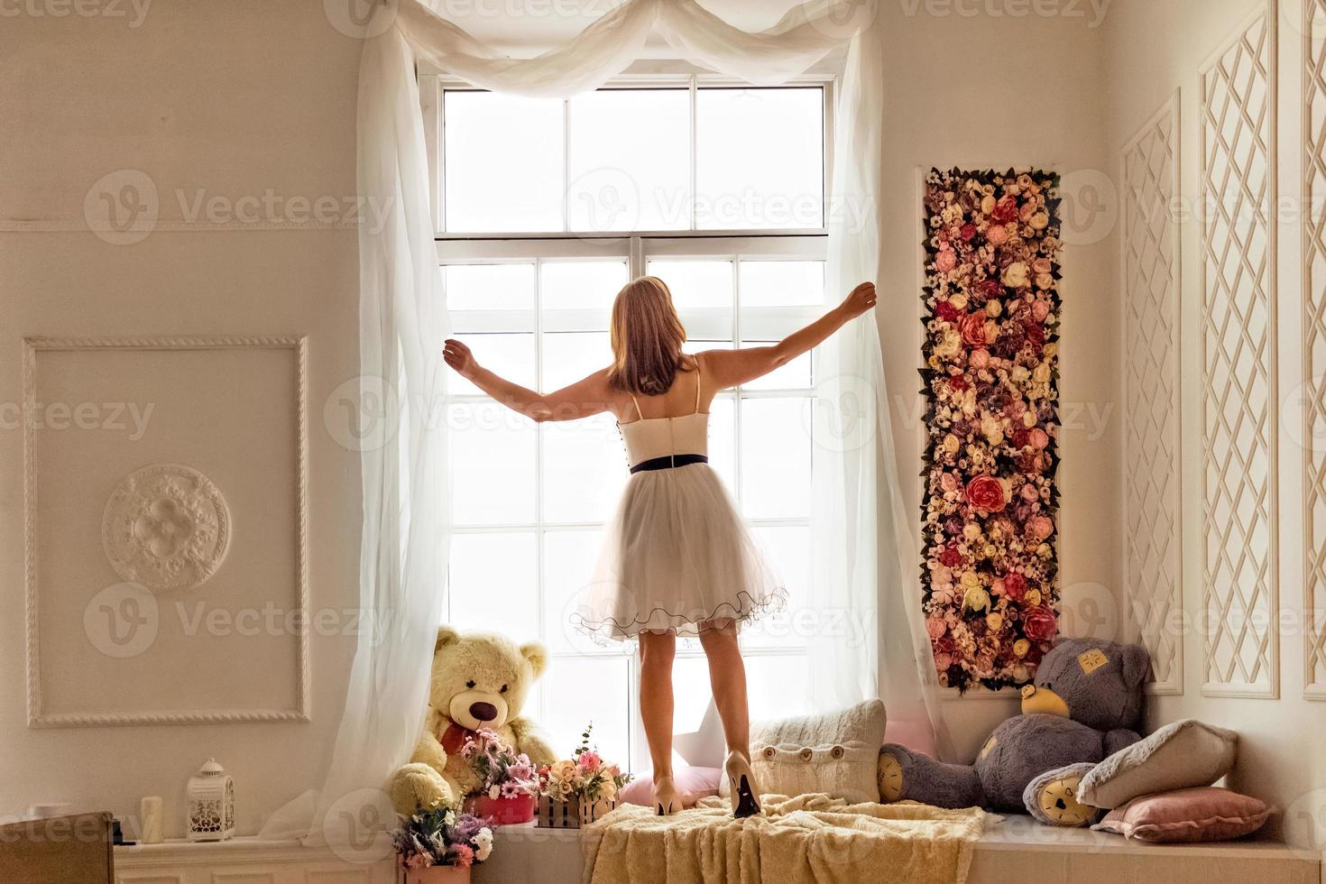 portrait d'une jeune femme vêtue d'une robe blanche redressant des rideaux blancs clairs près de la fenêtre. photo